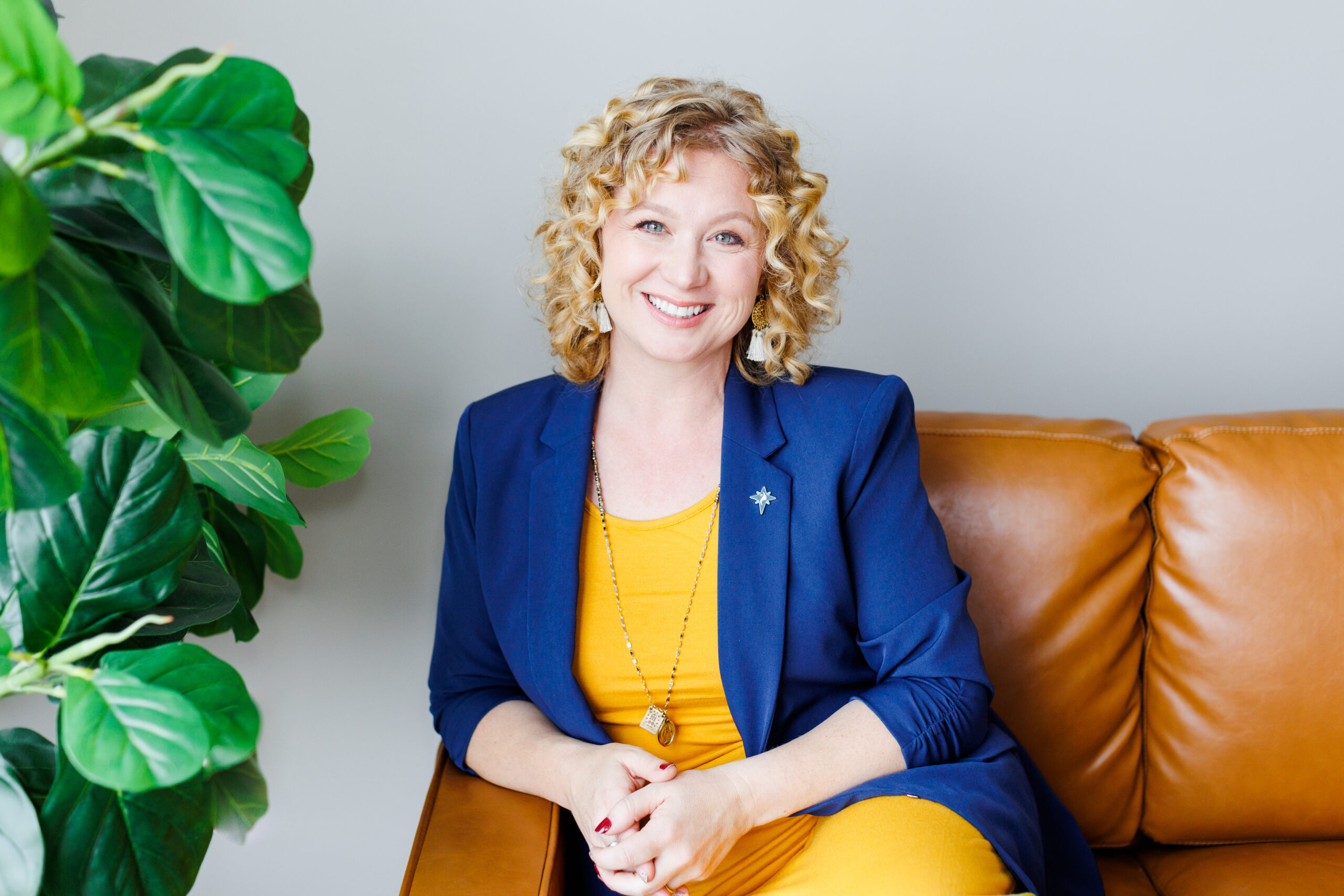 A blonde female in a yellow dress and bright blue blazer sits in a brown leather chair posing for a headshot. She is the founder of Guiding Star Project, a nationwide pregnancy resource center